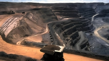 Open-pit mine from above showing stepped rock terraces, sandy slopes, and a haul truck on a foreground road